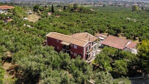 A large, red-brick building with multiple floors and balconies, surrounded by dense greenery and a hilly landscape in the background. - Villa San Andreas (Zakynthos)