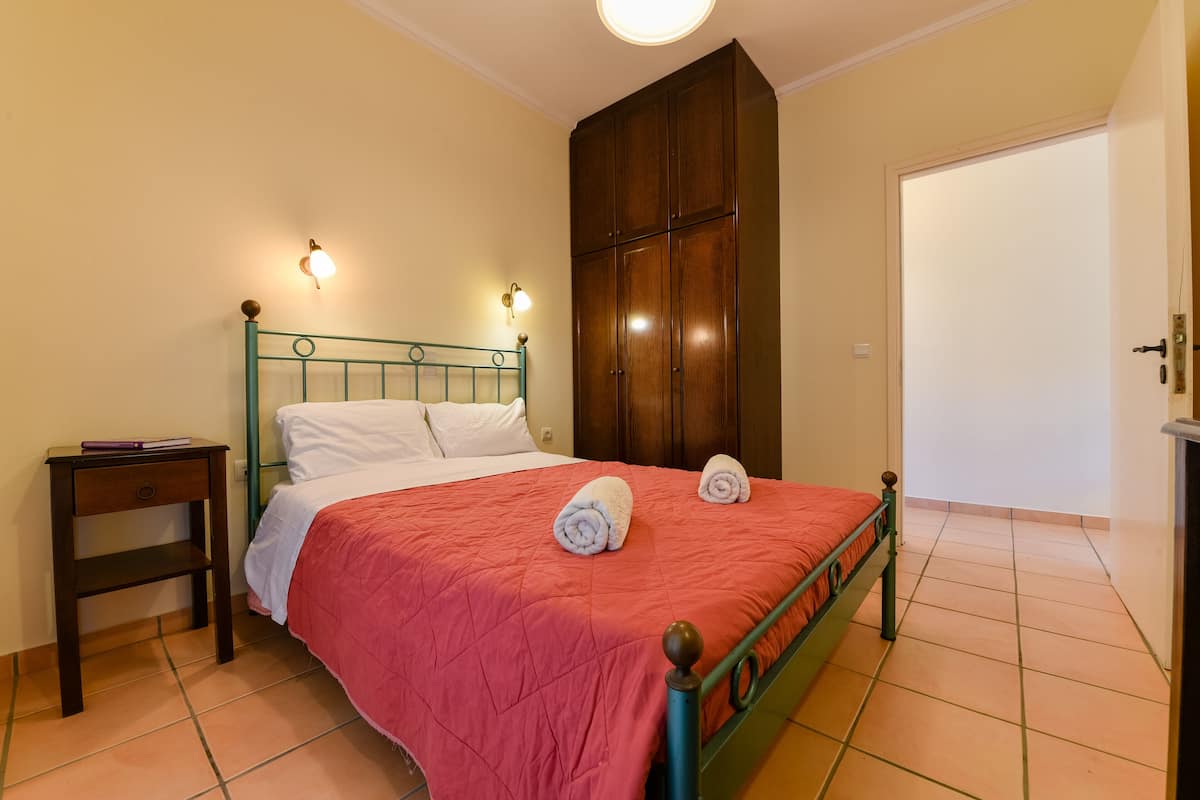 A neatly made bed with a red bedspread and two towels, a wooden wardrobe, a nightstand with a book, and a wall-mounted light fixture.