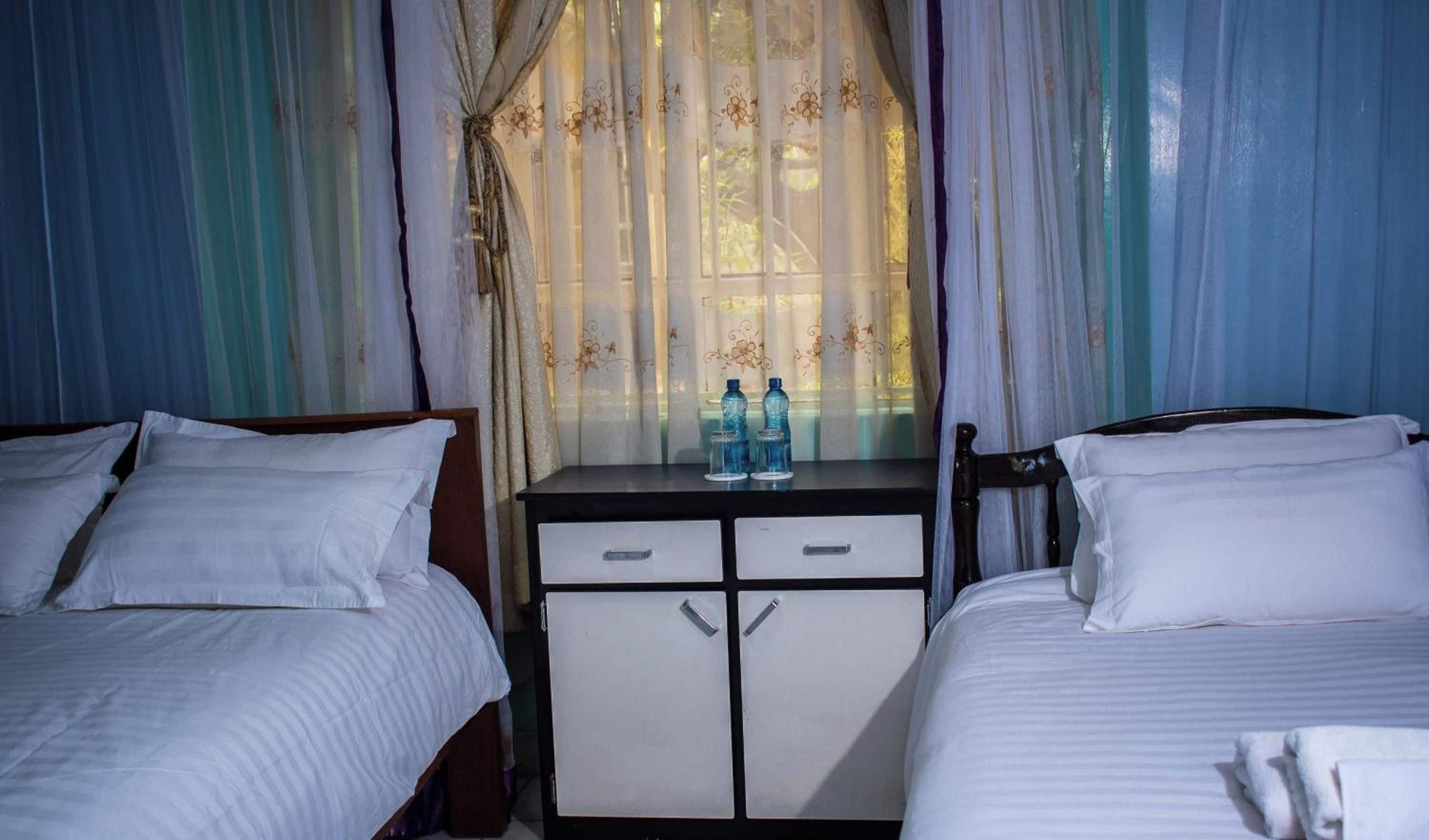 Habitación de hotel con dos camas, un escritorio pequeño con dos botellas de agua y una ventana con cortinas traslúcidas.
