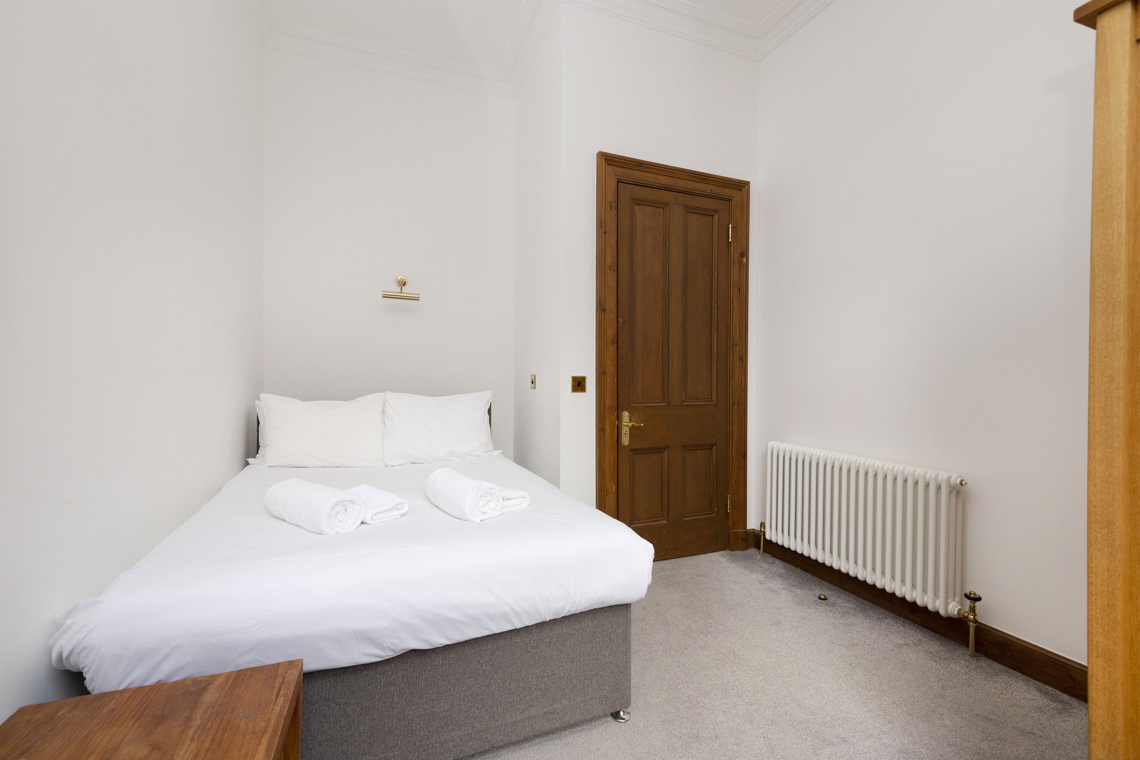 A neatly made bed with white linens and rolled towels, a wooden door, and a radiator on the floor.