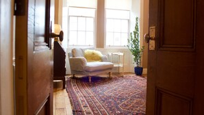 A hallway leading to a room with a large window, a white chair, a blue potted plant, and a patterned rug. - JOIVY Old Town Gem - Apartment With Great City Views (Edinburgh)