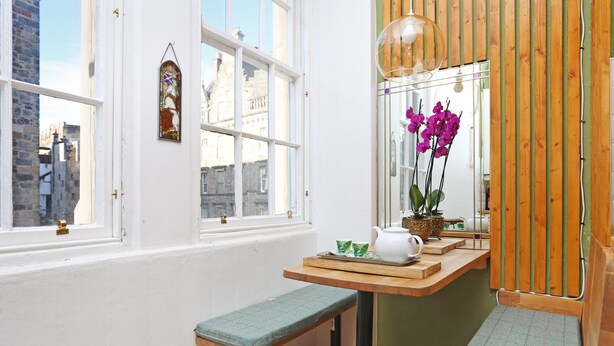 A cozy room with a wooden bench, a small table with a teapot, and a window with a view of buildings. - JOIVY Old Town Gem - Apartment With Great City Views (Edinburgh)