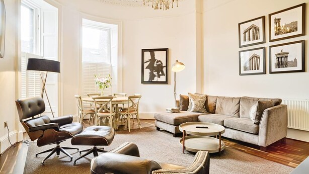 Un salon spacieux avec un canapé en cuir, une table à manger avec des chaises, un lustre et des tableaux encadrés accrochés au mur.