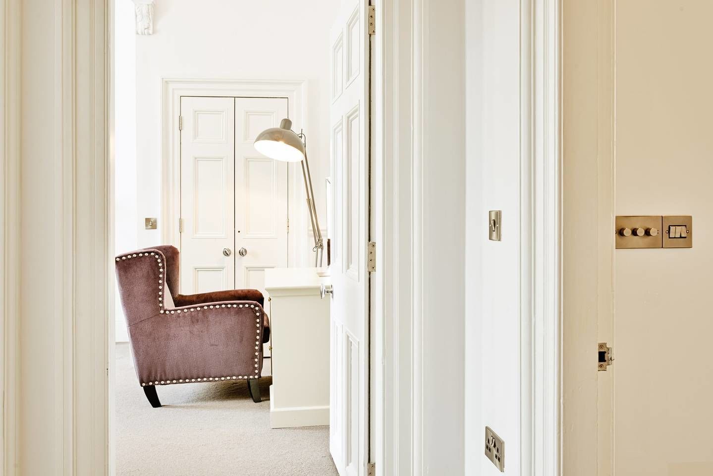 A hallway with a brown upholstered chair, a floor lamp, and a white door with a light switch and electrical outlets.