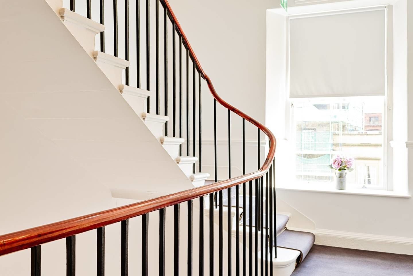A staircase with a wooden handrail and black balusters, leading to a room with a white door and a window with a view of buildings.