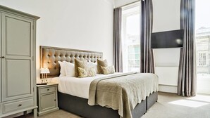 Une chambre à coucher avec une tête de lit capitonnée, un lit avec de nombreux oreillers, une table de chevet avec une lampe et une grande fenêtre avec des rideaux.