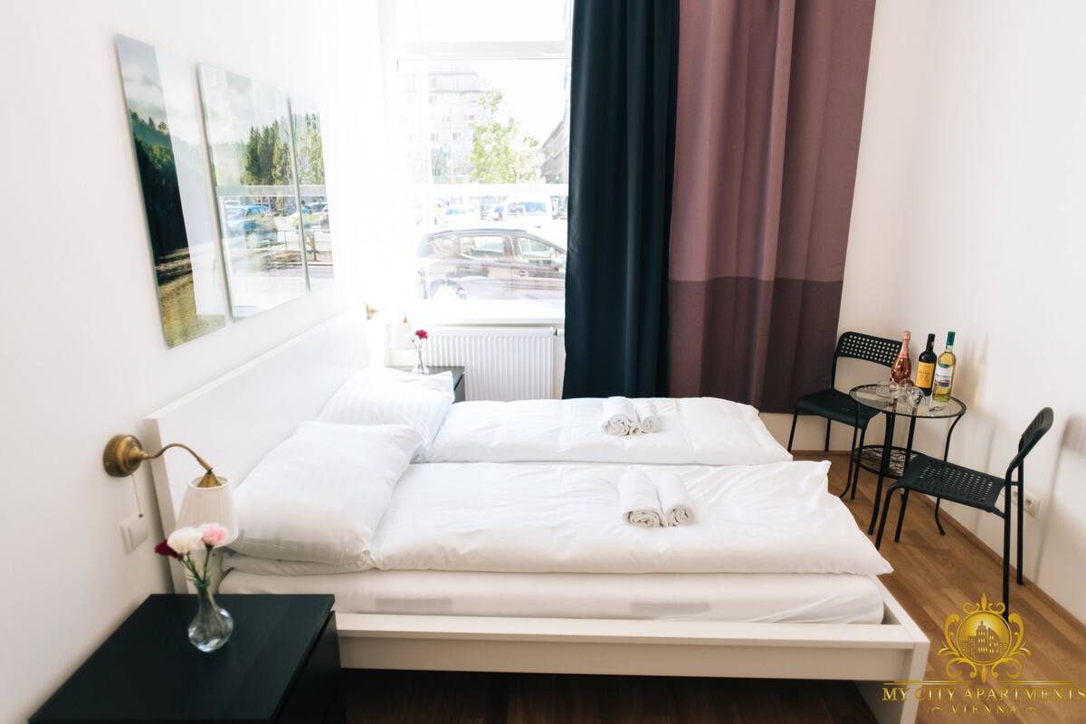 Une chambre d’hôtel avec un lit, une petite table sur laquelle sont posées des bouteilles, une chaise, un vase avec des fleurs et des œuvres d’art accrochées au mur.