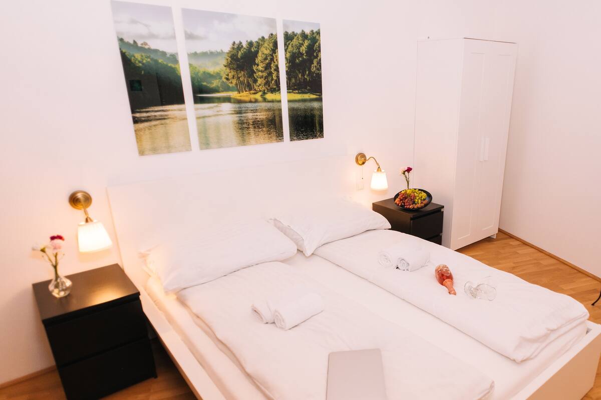 Un lit bien fait, avec du linge de lit blanc, une table de chevet avec une lampe et un tableau représentant un paysage accroché au mur.