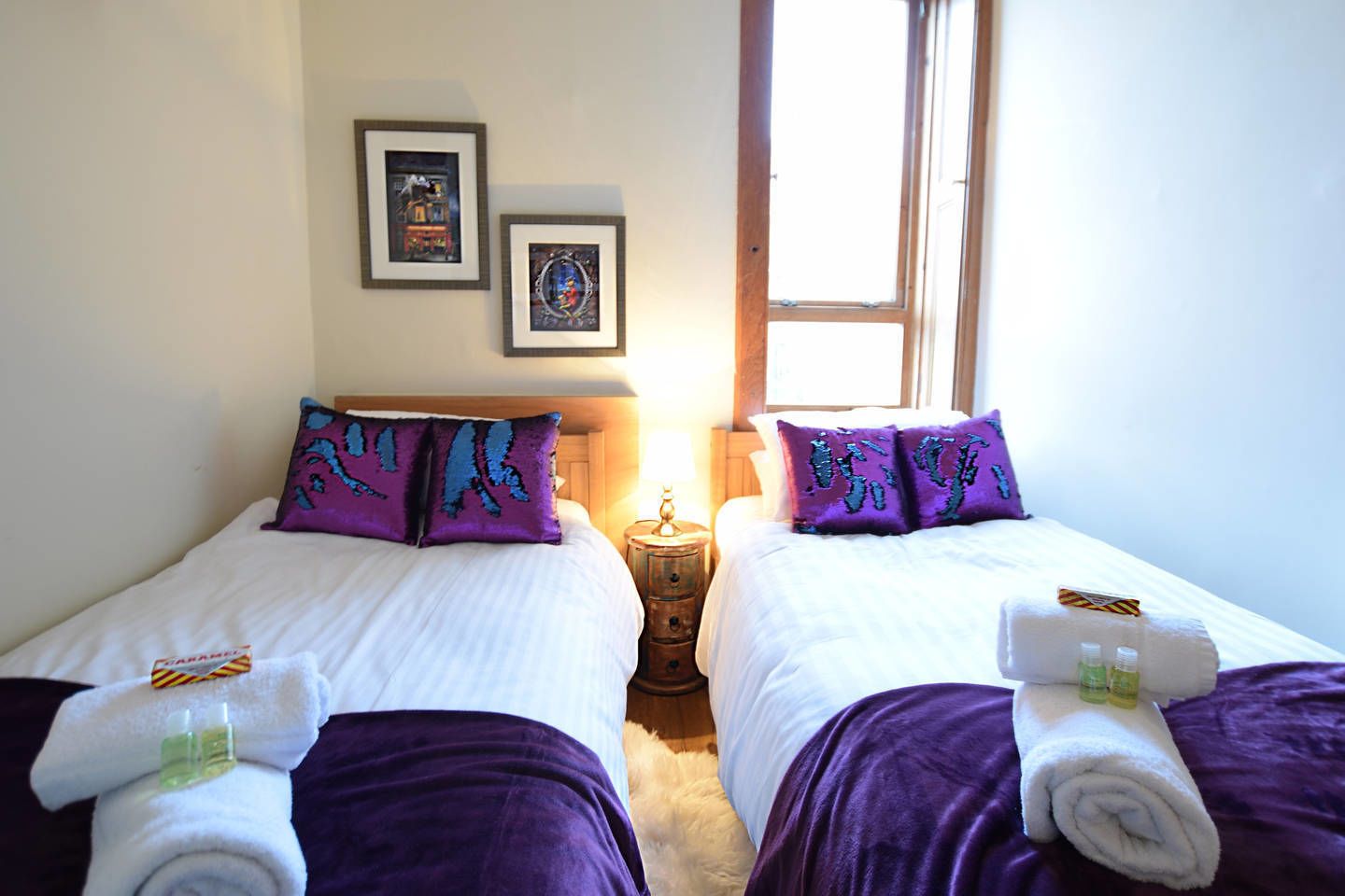 Ein Hotelzimmer mit zwei Betten, jeweils mit blau-lila gemusterten Kissen, weißer Bettwäsche und Handtüchern mit Toilettenartikeln. An der Wand befinden sich gerahmte Bilder und ein Fenster sorgt für natürliches Licht.