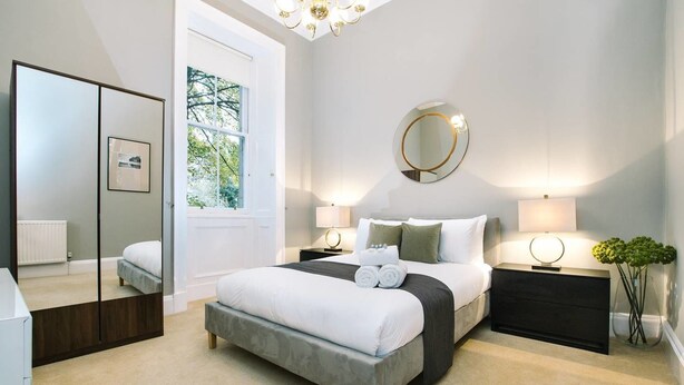 A bedroom with a large bed, bedside tables, a mirror, and a window with a view of trees. - JOIVY Palmerston Place Residence: Luxury City Centre Apt With Private Parking (Edinburgh)