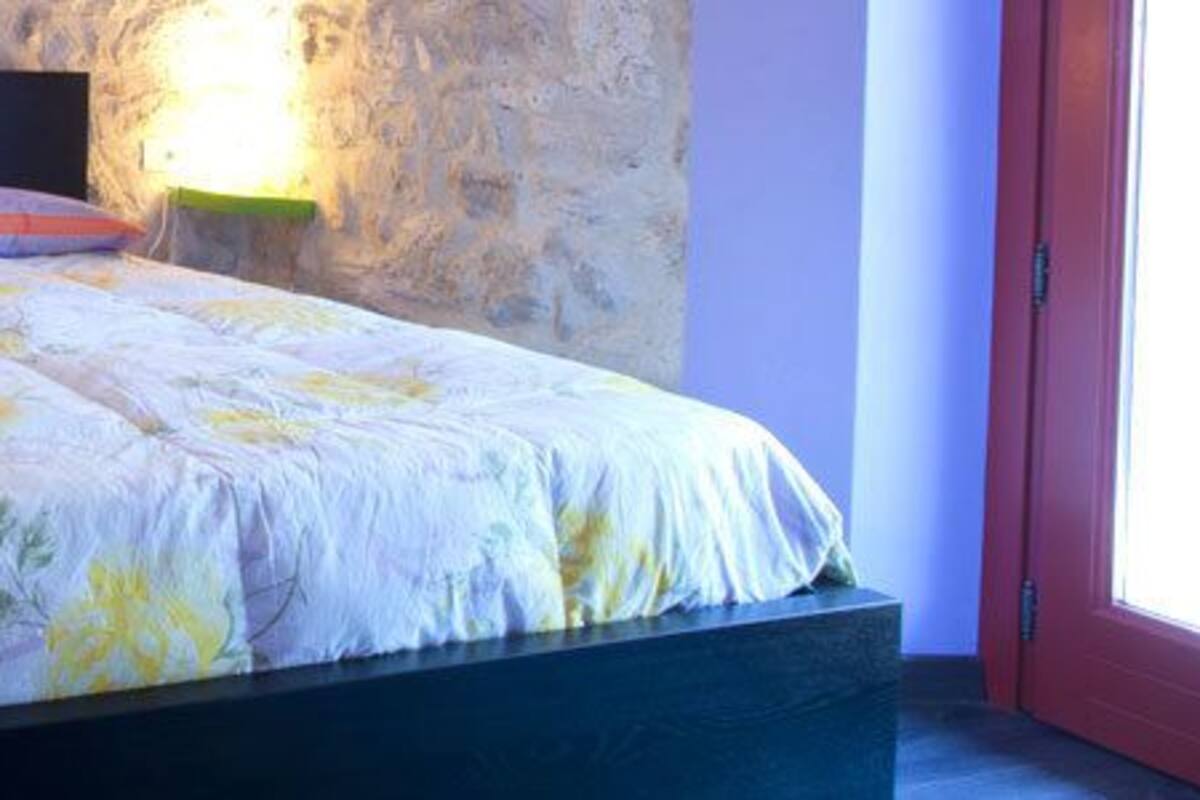 Una cama con colcha estampada, dos almohadas y lámparas de noche en una habitación con paredes de piedra y una puerta roja.