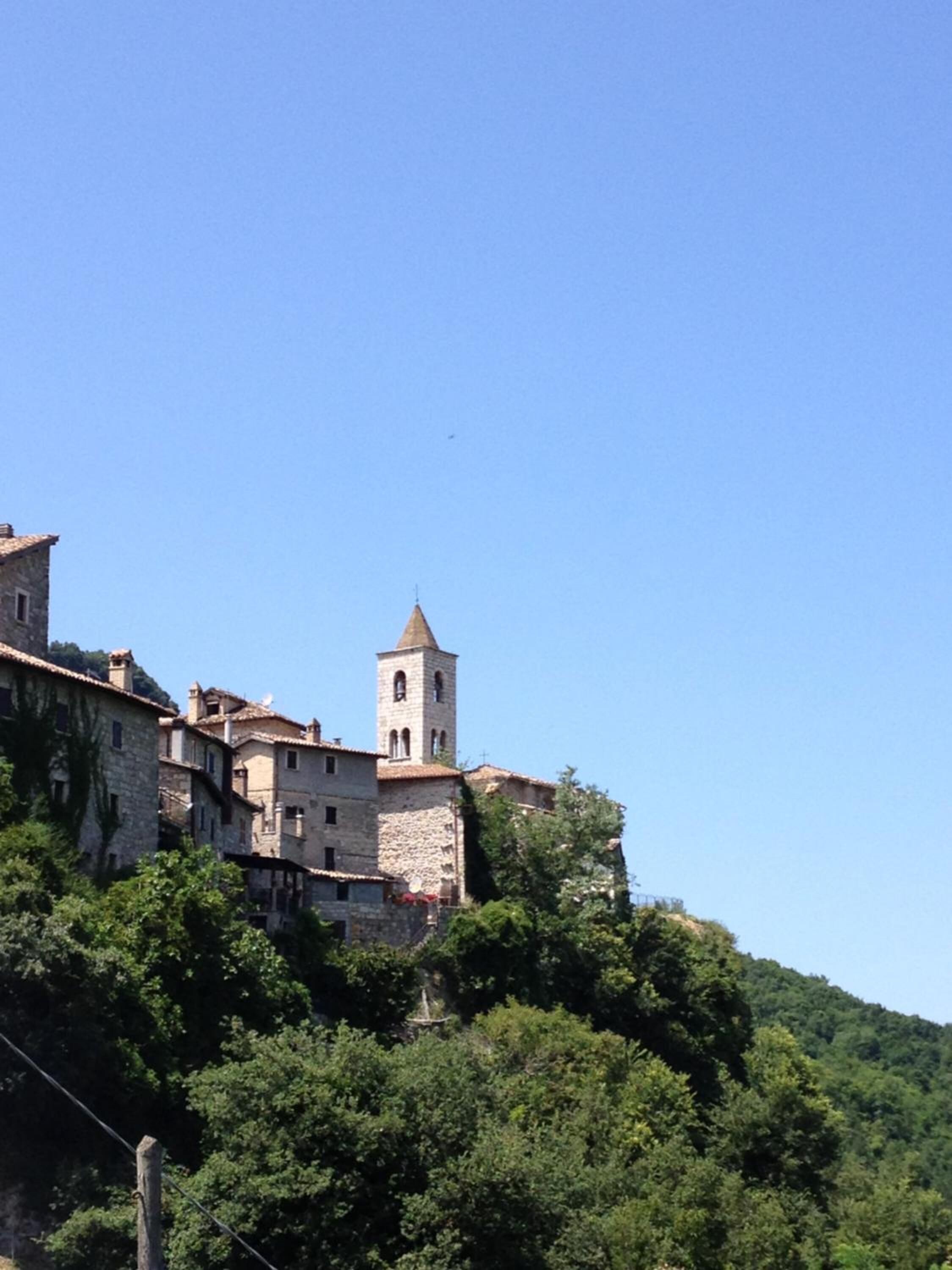 Un edificio storico in pietra con un campanile, circondato da un rigoglioso verde e alberi.