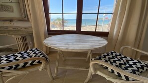 Une pièce avec une table ronde et deux chaises dotées de coussins à motifs, une grande fenêtre donnant sur la plage et un tableau encadré en bois accroché au mur.