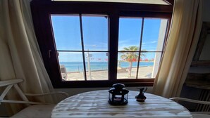 Une table ronde recouverte d’une nappe blanche, agrémentée de deux objets décoratifs : une lanterne et un pichet. La table est placée devant une fenêtre donnant sur la plage et les palmiers.