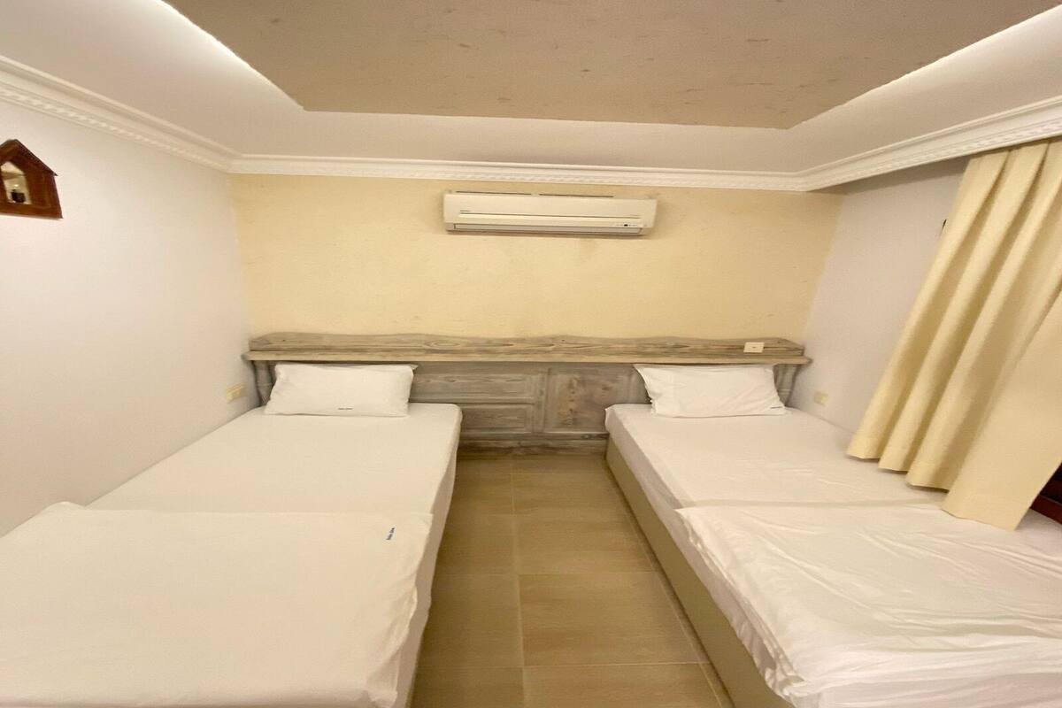 A room with two single beds, a wooden headboard, an air conditioning unit, and a curtain on the right side.