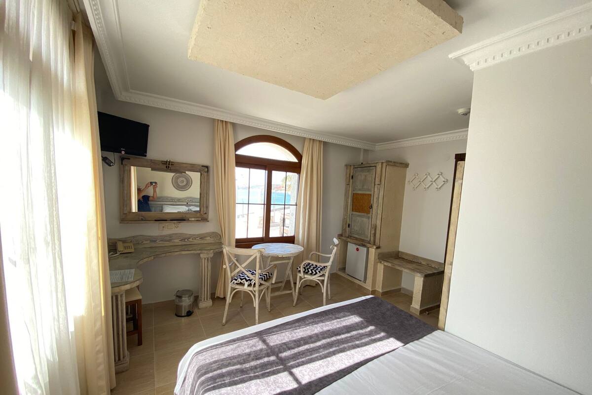 A hotel room with a bed, a desk, a chair, a small fridge, a mirror, and a window with curtains.