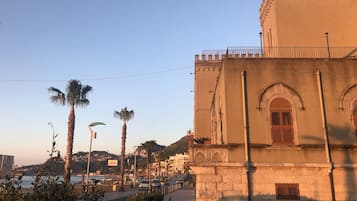 Un edificio frente al mar con ventanas arqueadas y un balcón, con vistas a una calle con palmeras y coches estacionados.