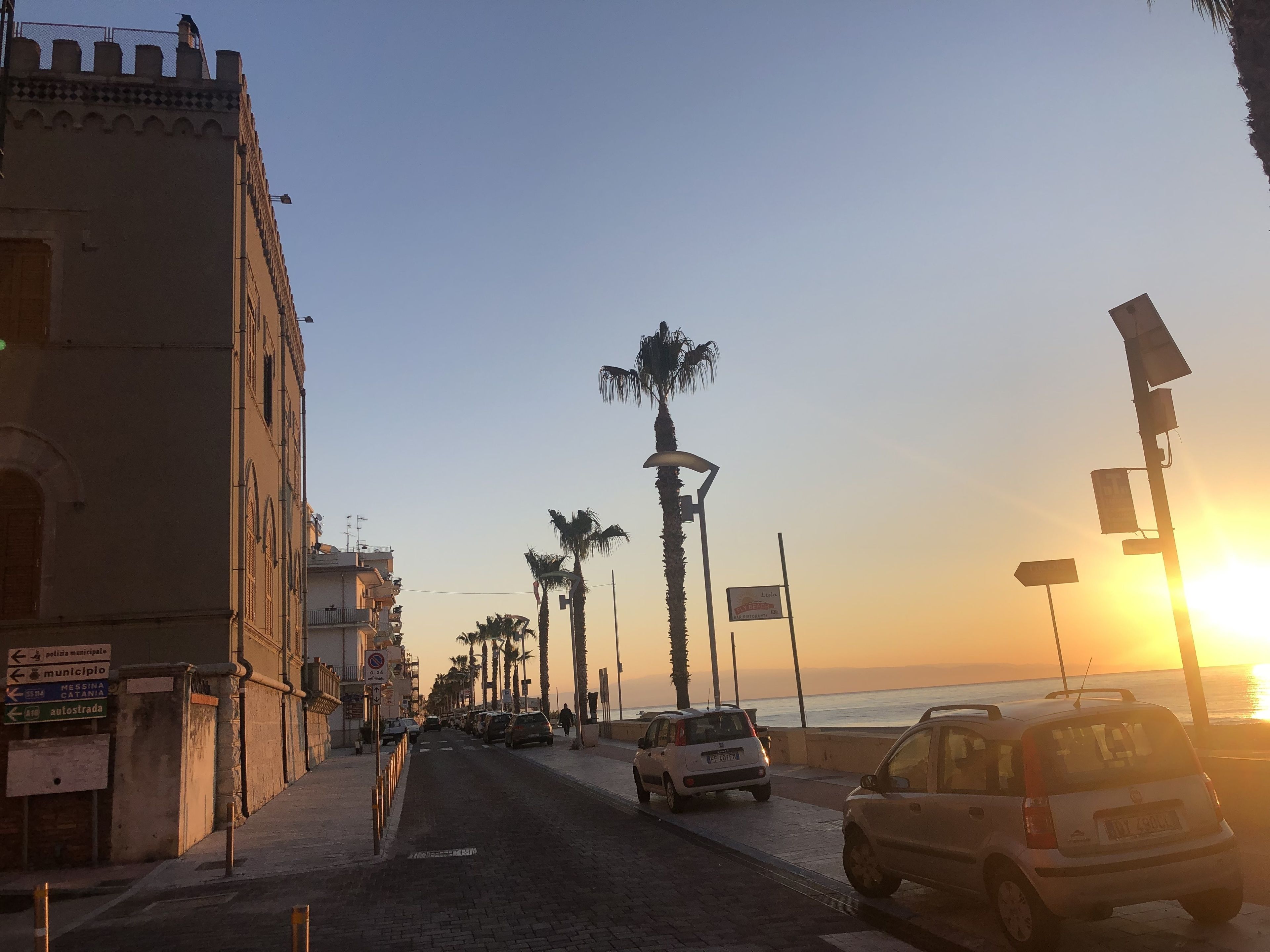 A coastal street with parked cars, palm trees, and a sunset.