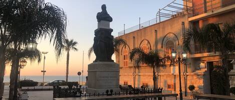 A statue in a plaza with benches and palm trees, surrounded by buildings with balconies and arched windows.