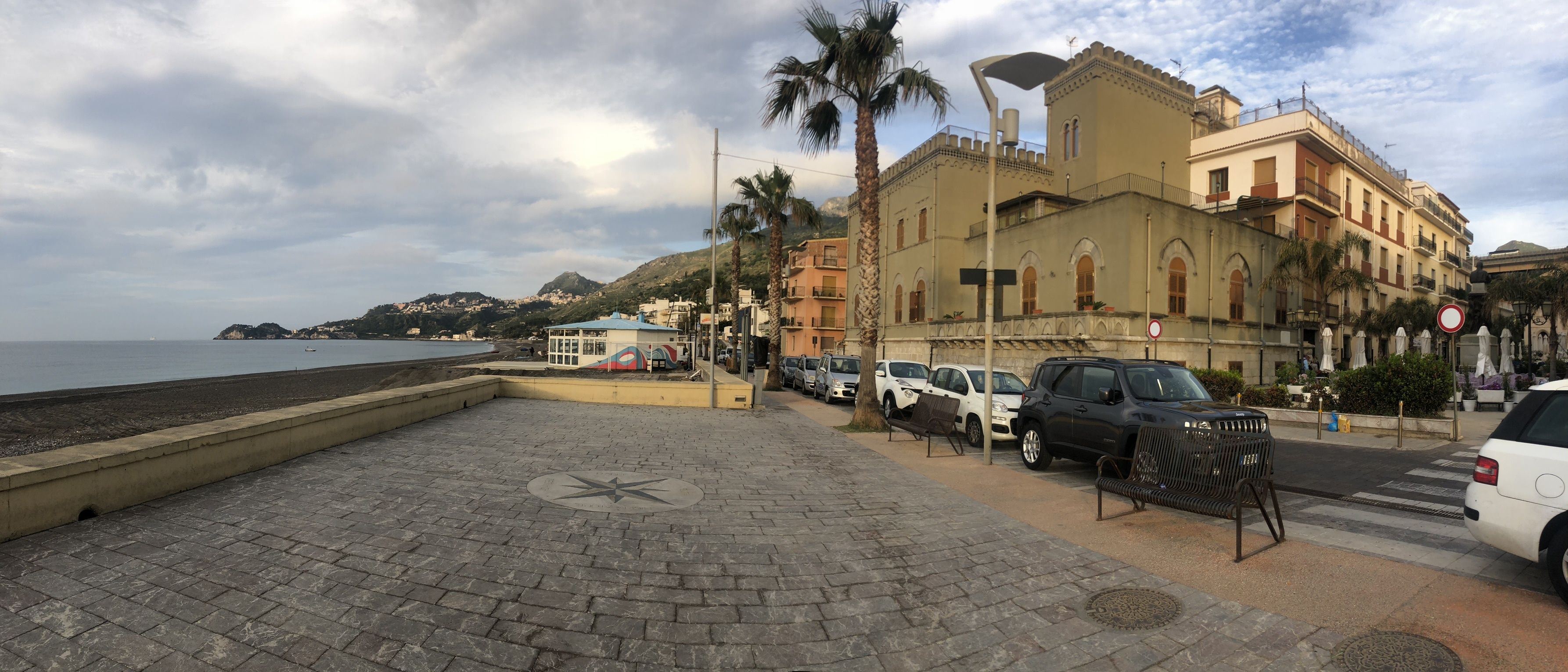 A paved waterfront promenade with a palm tree, parked cars, and a building with arched windows.