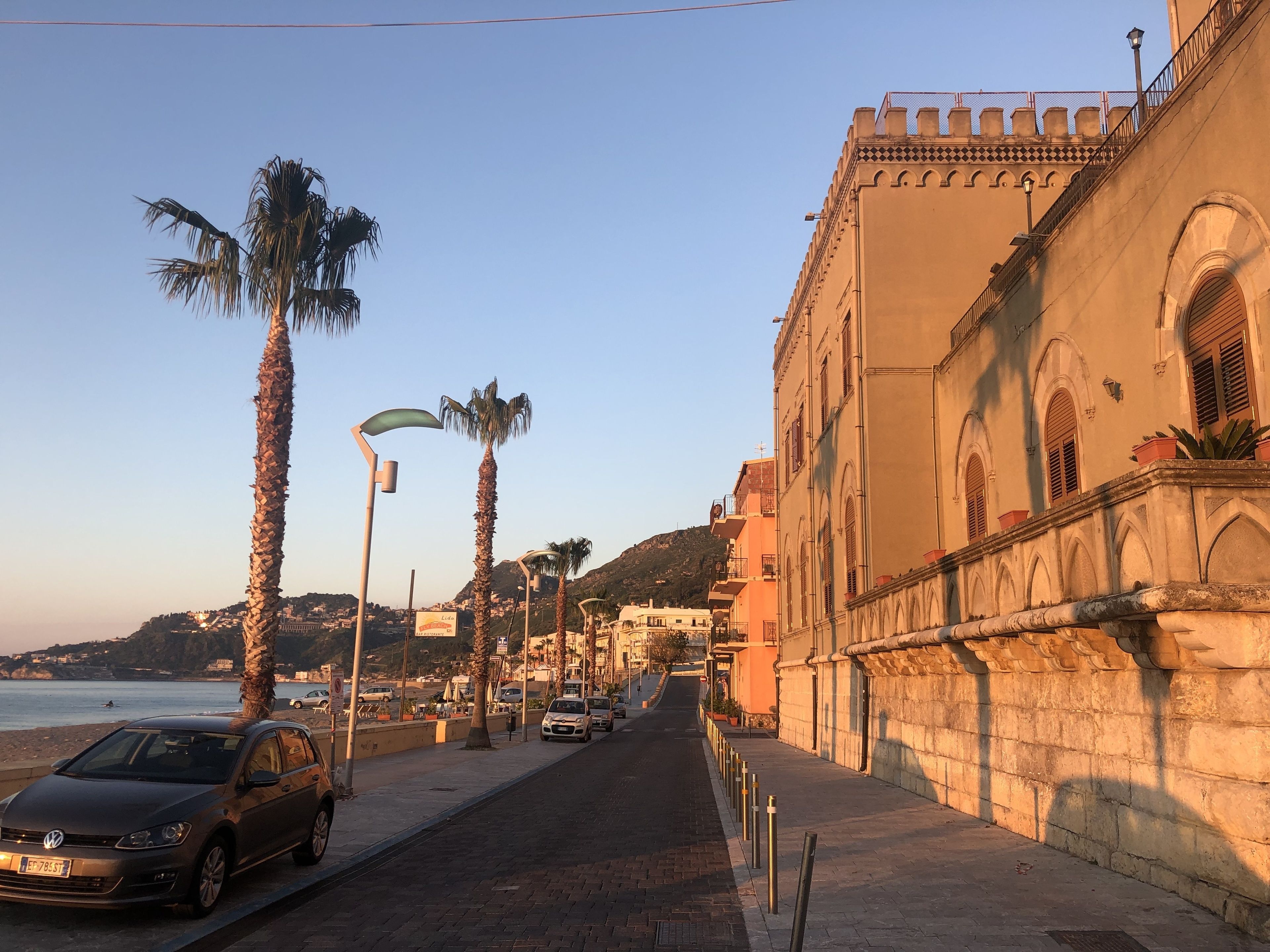 A street with palm trees, a parked car, and a building with arched windows and a balcony.