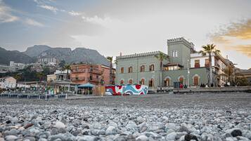 A coastal town with a prominent building featuring a tower and arched windows, a pebbled beach, and a backdrop of mountains.