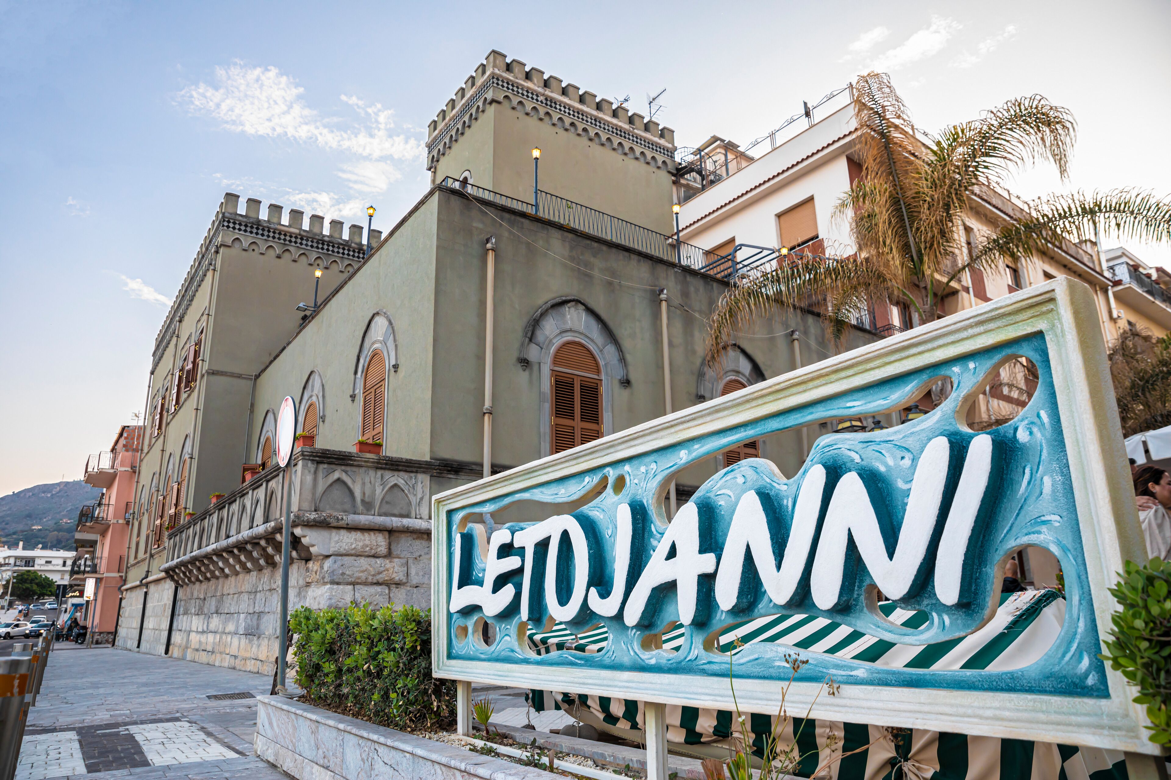 A sign with the name "LETOJANNI" in front of a building with classical architecture.