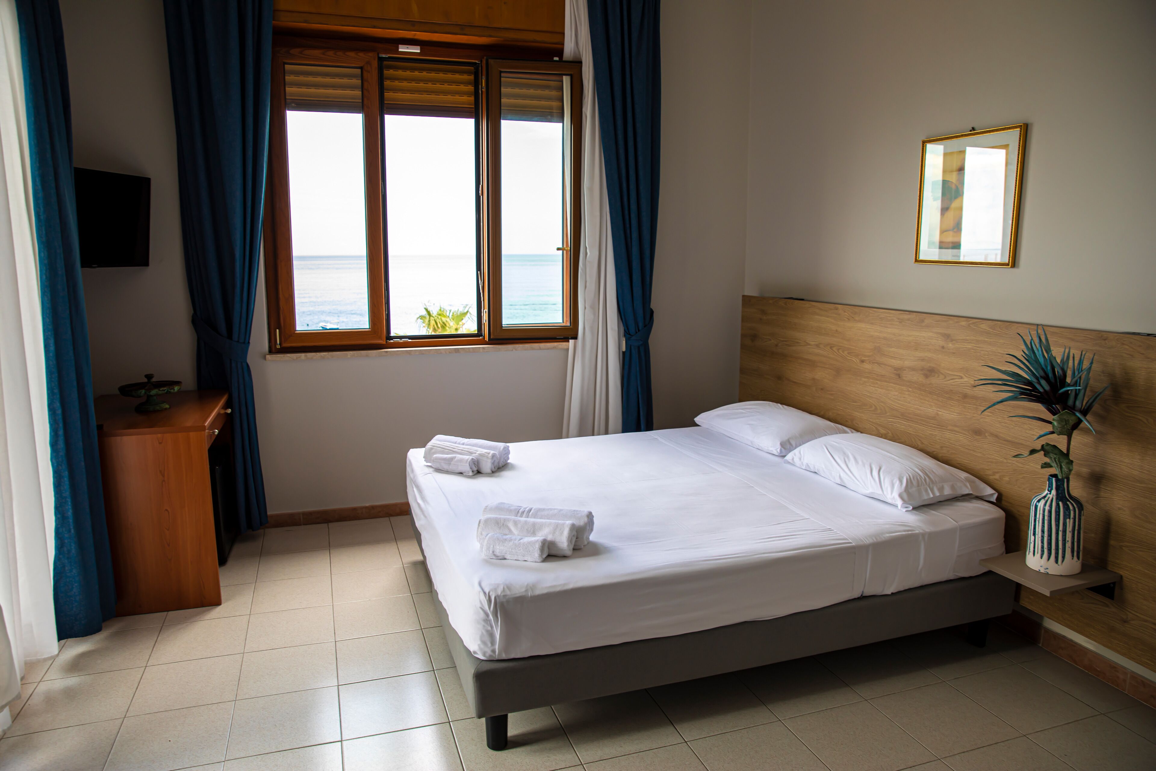 A neatly made bed with white linens and towels, a wooden headboard, a bedside table with a plant, and a window with curtains.