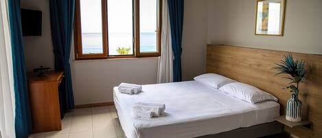 A neatly made bed with white linens and towels, a wooden headboard, a bedside table with a plant, and a window with curtains.