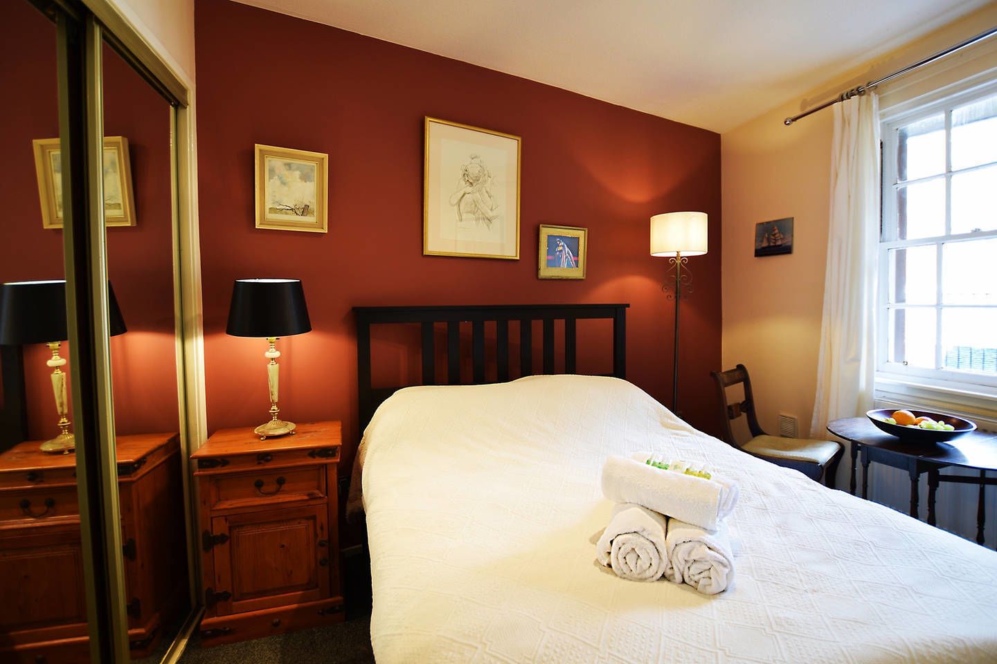 A hotel room with a large bed, a nightstand with a lamp, a chair, a small table with fruit, and framed artwork on the walls.