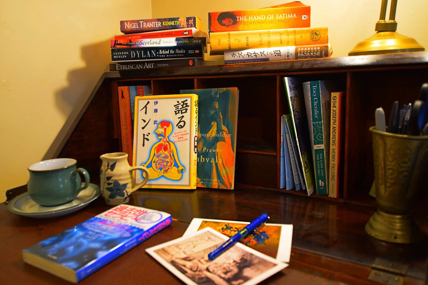A desk with a stack of books, a cup, a mug, a pen, and a small vase.