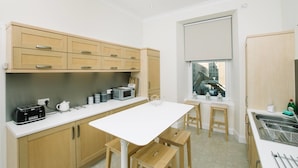 A modern kitchen with wooden cabinets, a white countertop, a dining table with benches, and a window with a city view. - JOIVY Great Location! Heart of City Centre 3bed Apartment (Edinburgh)