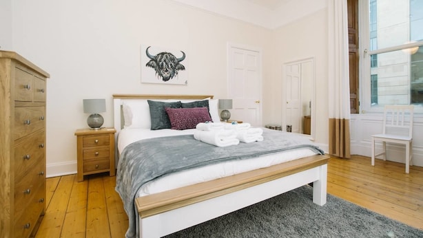 A bedroom with a wooden bed, a grey blanket, a bull artwork on the wall, and a wooden dresser. - JOIVY Great Location! Heart of City Centre 3bed Apartment (Edinburgh)
