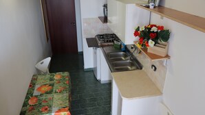 A compact kitchen with a gas stove, sink, and a small countertop. There is a table with a floral tablecloth and a chair. A decorative plant is on the countertop. - Casa Vacanza Villa Sole (Siculiana)