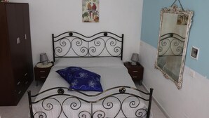 A bedroom with a metal bed frame, a wooden wardrobe, a wall-mounted mirror, and a small bedside table with a lamp. - Casa Vacanza Villa Sole (Siculiana)