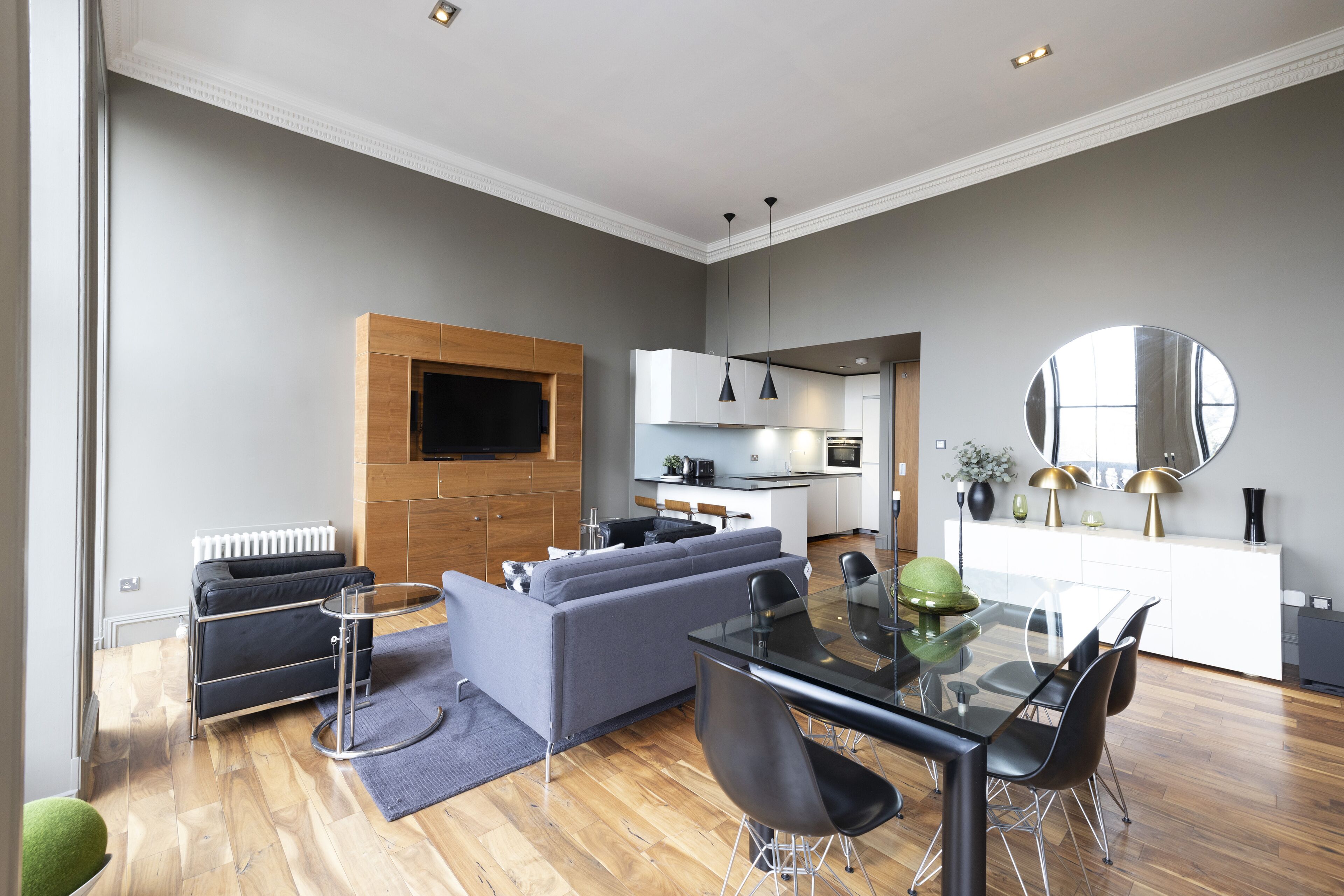 A modern living room with a grey sofa, a glass dining table, and black chairs. There is a kitchen in the background with white cabinets and a large mirror above the counter.