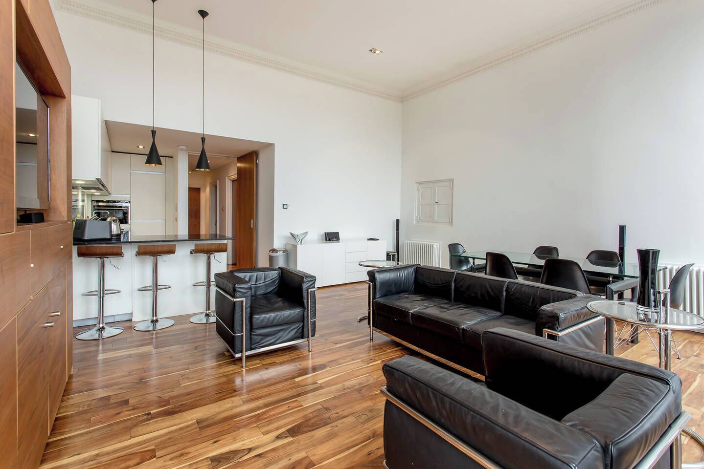Un salon moderne avec un plancher en bois, un canapé en cuir noir, un fauteuil en cuir noir et une table à manger en verre avec des chaises.