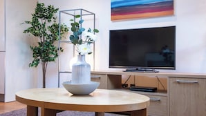 Ein modernes Wohnzimmer mit einem runden Holztisch, einer weißen Vase mit Blumen, einem großen Flachbildfernseher und einer schlichten Wandregal-Einheit.