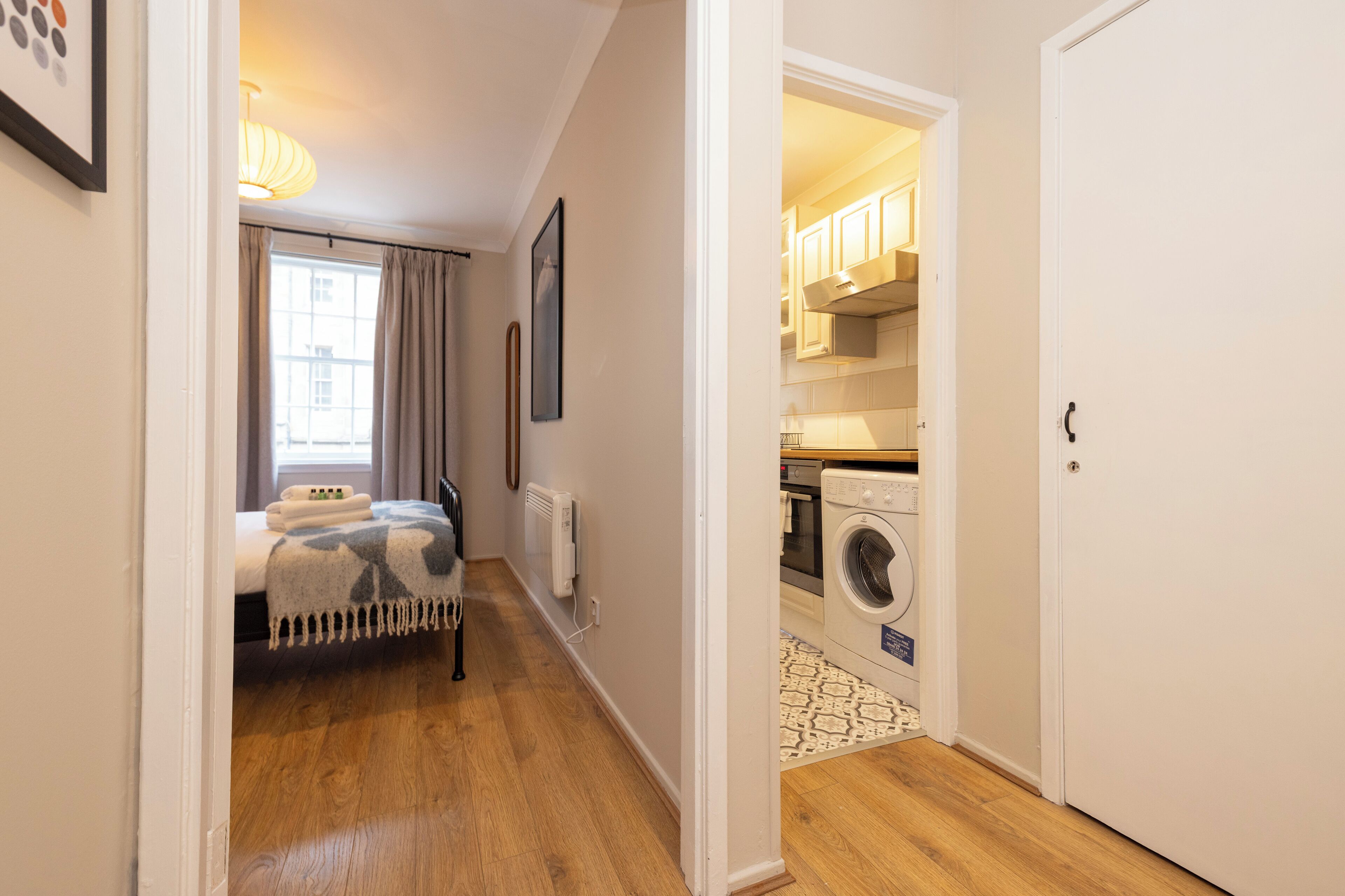 A compact, well-lit room with a bed, a radiator, and a washing machine in the adjacent space.