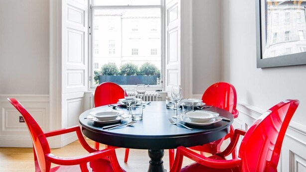 A dining area with a round table set for six, red chairs, and a view of buildings through large windows. - JOIVY York Place Apartment in Georgian City Centre Building (Edinburgh)
