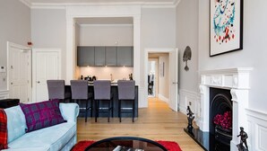 A modern living room with a light blue sofa, a red rug, and a glass coffee table. There is a kitchen in the background with grey cabinets and a dining area with bar stools. A fireplace with a statue is visible on the right. - JOIVY York Place Apartment in Georgian City Centre Building (Edinburgh)