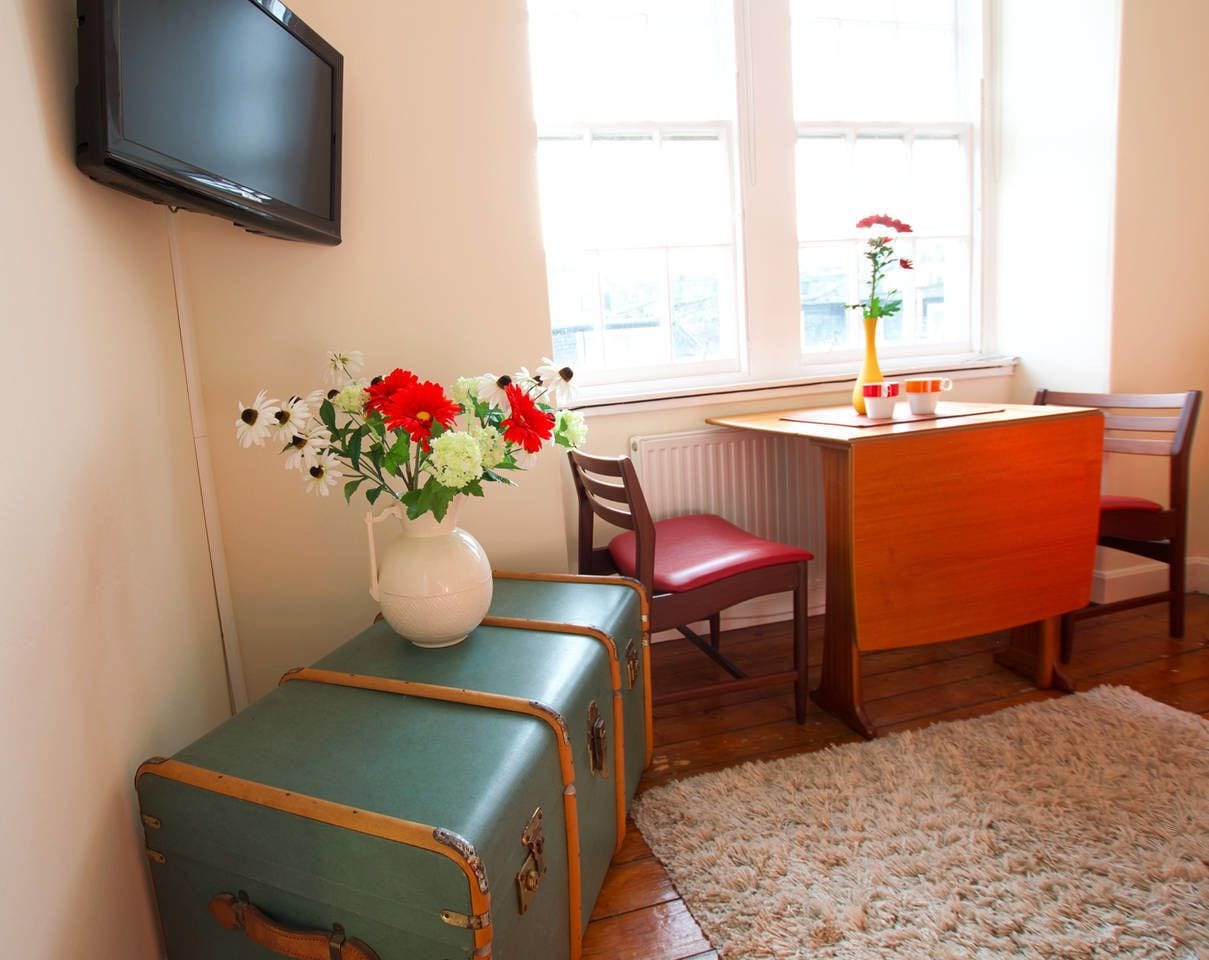 Een kamer met een houten tafel, twee vintage koffers, een bloemenvaas, een rode stoel en een aan de muur bevestigde televisie.