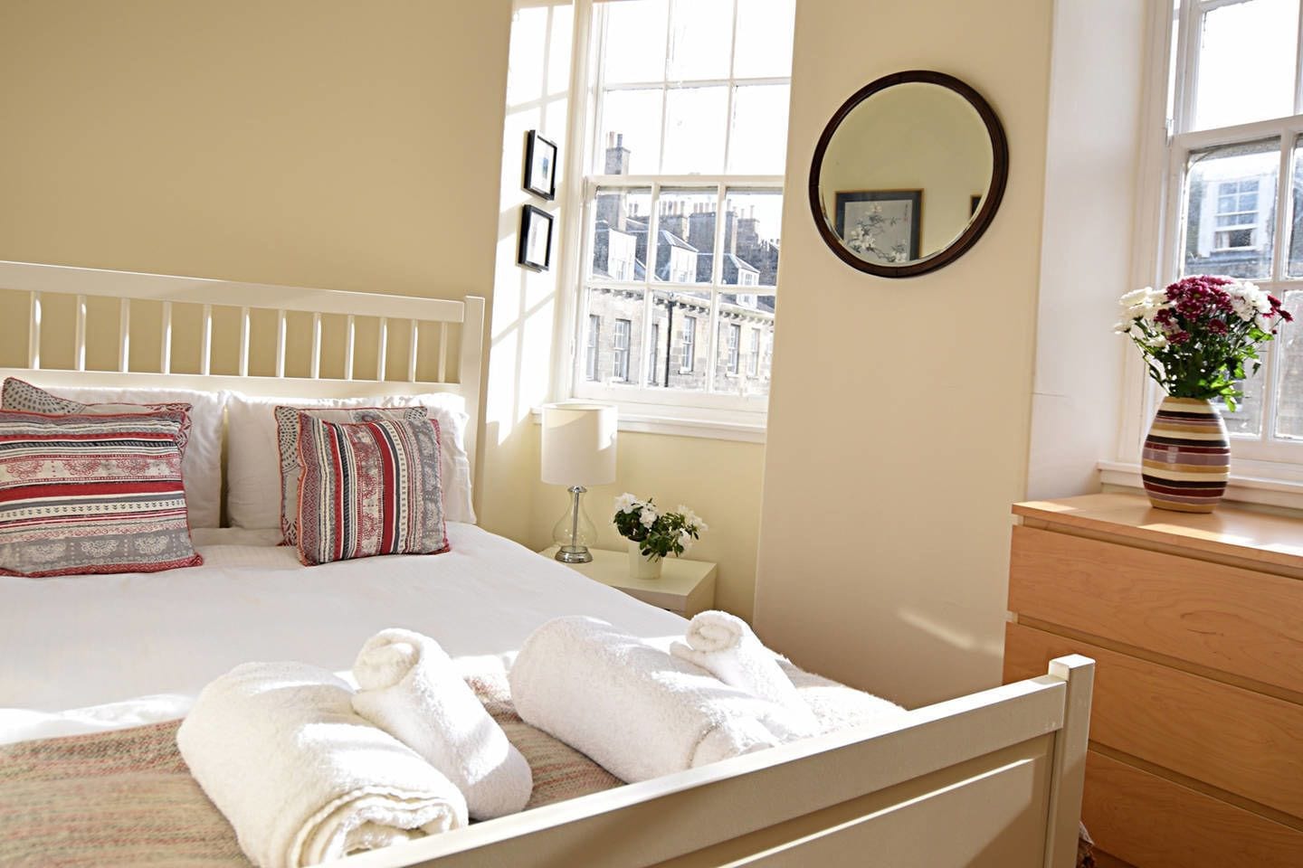A neatly made bed with pillows, a bedside table with a lamp, a dresser, and a window with a view of buildings.
