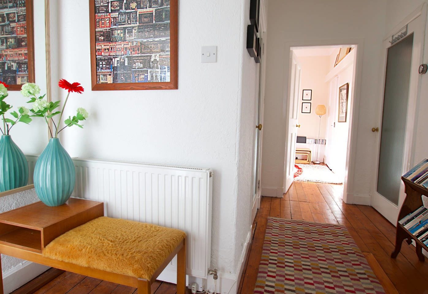 A room with a wooden bench, two teal vases with flowers, a white radiator, a patterned rug, and a bookshelf with books.