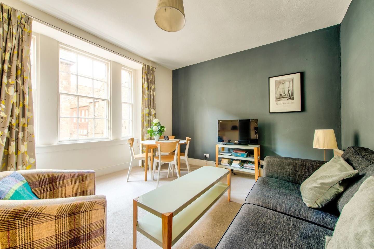 A living room with a sofa, a coffee table, a TV stand with a flat-screen TV, a dining table with chairs, and a window with curtains.