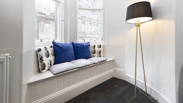 A room with a bench by a window, a floor lamp, and a view of a building outside. - JOIVY Iconic Cockburn Street 4 Bedroom Apartment: Heart of Old Town (Edinburgh)