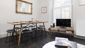 A modern living room with a wooden dining table, black stools, a large flat-screen TV, and a window with blinds. - JOIVY Iconic Cockburn Street 4 Bedroom Apartment: Heart of Old Town (Edinburgh)