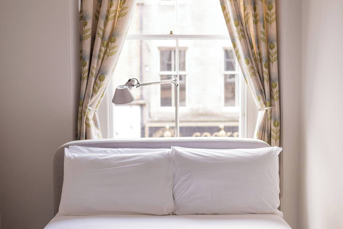 A bed with white linens and a bedside lamp, curtains with a floral pattern, and a view of a building through the window.