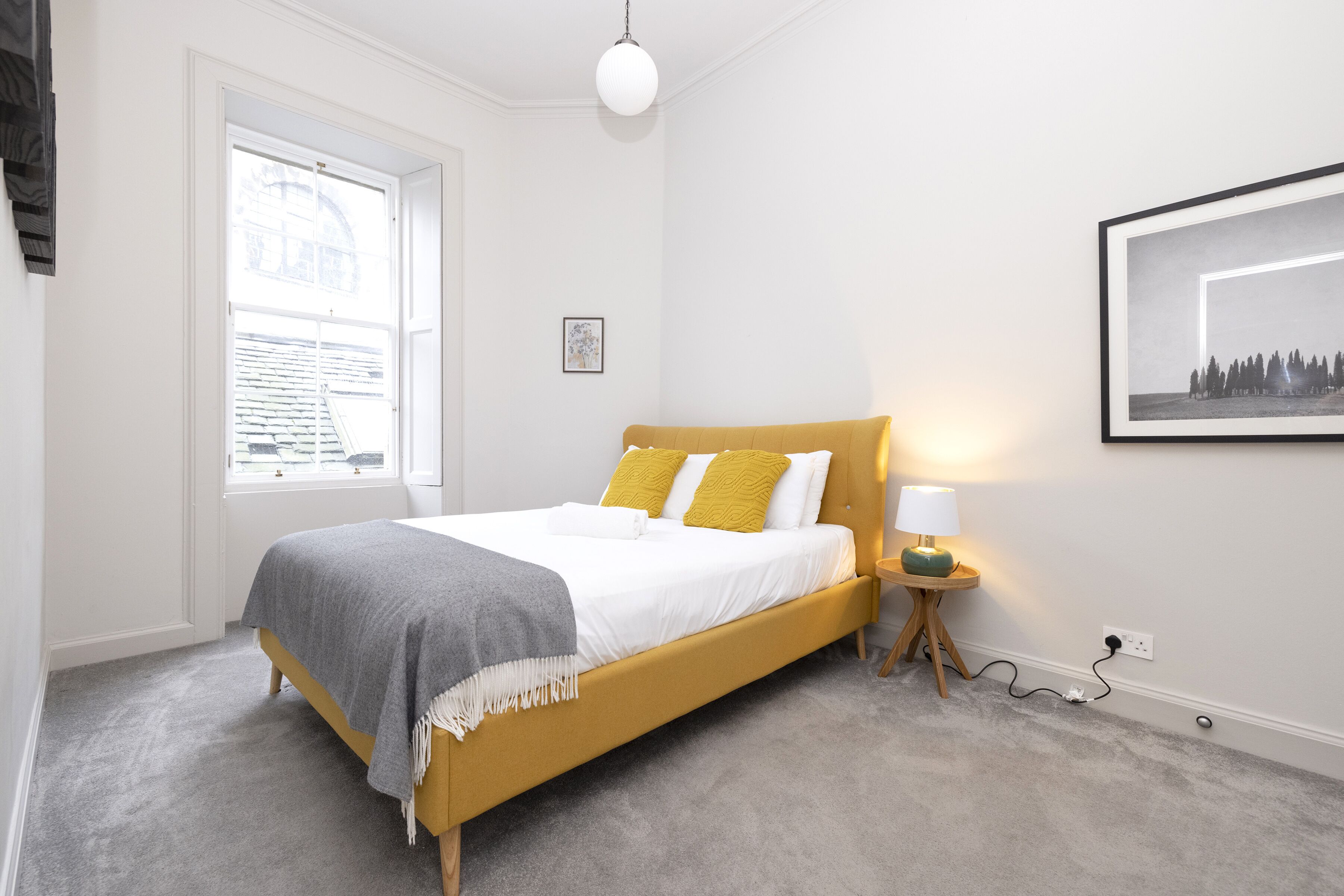 Ein Schlafzimmer mit einem Bett, einem Nachttisch mit Lampe, einem Fenster und einem gerahmten Bild an der Wand.