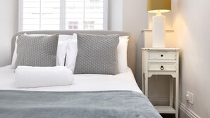 A neatly made bed with a grey blanket and pillows, a bedside table with a lamp, and a window with blinds. - JOIVY Iconic Cockburn Street 4 Bedroom Apartment: Heart of Old Town (Edinburgh)